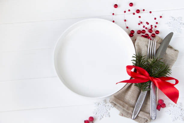 istock Christmas table setting-white plate, table fork and knife set with napkin, fir branch and red ribbon, white snowflakes, red berries on white wooden board 1343468732