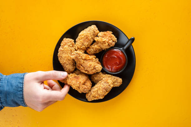 Crispy fried chicken wings with tomato sauce Crispy fried chicken wings with tomato sauce on plate, yellow background. Male hand picking chicken wing, junk food, unhealthy eating concept. Top view copy space hand thigh stock pictures, royalty-free photos & images