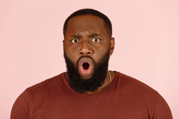 Funny shocked African-American guy actor on pink background Funny shocked wide-eyed African-American guy actor with open moth in t-shirt poses for camera on pink background in studio extreme close view mesmerised stock pictures, royalty-free photos & images
