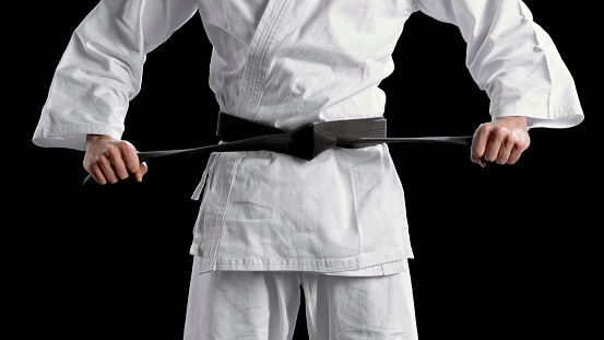 Close-up of young male karate player tightening his black belt while standing against black background.