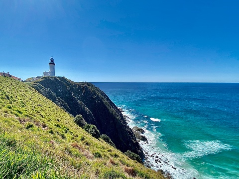 horizontal seascape of white lighthouse on clear sky day with green grass headland and rocks going out to blue ocean at Australia’s most easterly point at Byron Bay NSW Australia