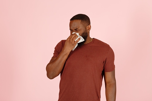 Sick African-American man with nasal cold blows his nose at white tissue standing on light pink background in studio closeup