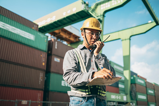 Dock worker standing against cargo containers having conversation with loading control room for shipping plans