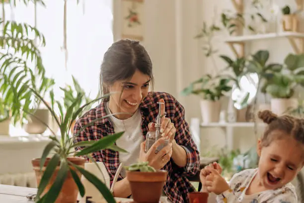 Photo of Young happy mixed race woman with a little daughter is planting houseplants at home and having fun splashing water.Home gardening.Family leisure,hobby concept.Biophilia design and urban jungle concept