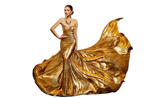 Woman in Long Golden Evening Dress waving in Air. Elegant Fashion Model in Shiny Luxury Holiday Gown with Gold Jewelry over Studio White Background