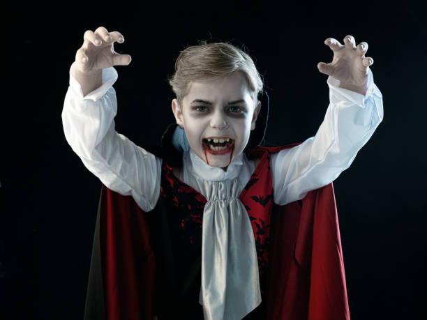 Boy in Halloween vampire makeup costume Portrait of boy wearing Halloween vampire makeup and costume cloak bared his teeth, isolated on black background. vampire stock pictures, royalty-free photos & images