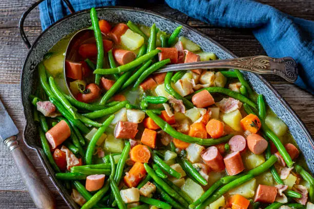 Traditional and rustic stew or soup with green beans, vegetables and potatoes, Cooked with bacon and and served with vienna sausage in a pot with ladle on rustic and wooden table. Closeup and isolated view from  above