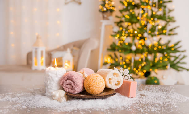istock Winter spa pampering concept. Various self care products bath bomb, soap bar, luffa sponge on wood tray with candle burning and Christmas tree with lanterns on background. 1343500151