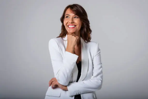 Studio portrait shot of attractive middle aged woman with toothy smile wearing blazer while standing at isolated grey background. Copy space.