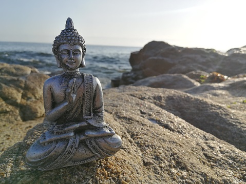 Silver Buddha statue on rocks at the beach, some of the rocks and the sea are out of focus, sunny day with a very clear sky, the rocks are wet and seaweeds can be seen out of focus in the back, with copy space, horizontal photo, ideal for meditation and relaxation, alternative medicine, spa background