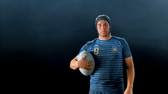 Portrait of male rugby player standing with ball against black background.