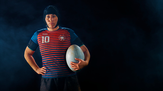 Portrait of female rugby player standing with ball against black background.