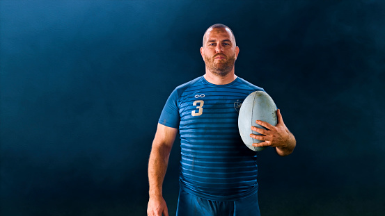 Portrait of male rugby player standing with ball against black background.