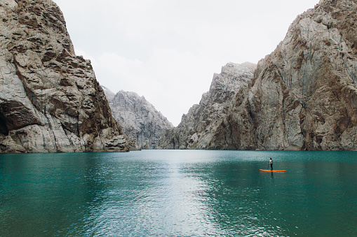 Woman traveler surfing by the orange SUP board at the picturesque crystal blue lake Kol-Suu surrounded by the high mountain peaks in Kyrgyzstan
