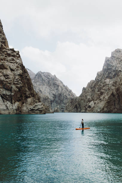 Woman paddleboarding at the scenic remote turquoise mountain lake in Tian Shan, Central Asia Woman traveler surfing by the orange SUP board at the picturesque crystal blue lake Kol-Suu surrounded by the high mountain peaks in Kyrgyzstan kyrgyzstan photos stock pictures, royalty-free photos & images