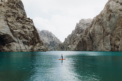 Woman traveler surfing by the orange SUP board at the picturesque crystal blue lake Kol-Suu surrounded by the high mountain peaks in Kyrgyzstan