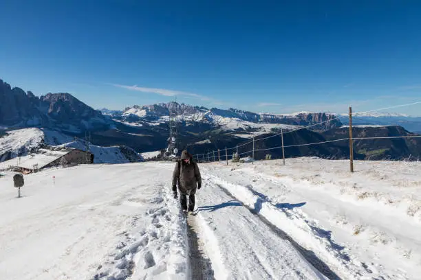 Photo of A woman in a warm winter jacket with photo gear goes down a snowy path, in clear sunny weather, to the top of Seceda Mountain in the Italian Dolomites. In the mountains is snow and blue sky with cloud.