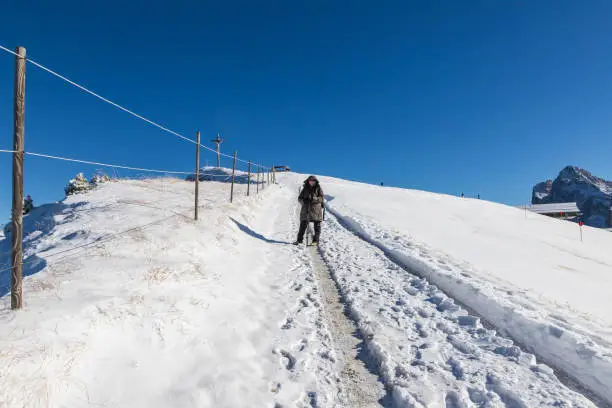 Photo of A woman in a warm winter jacket with photo gear goes down a snowy path, in clear sunny weather, to the top of Seceda Mountain in the Italian Dolomites. In the mountains is snow and blue sky with cloud.