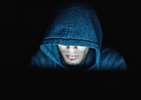Ominous-looking man in hoodie stares down at computer: hacker or cyber-criminal perhaps