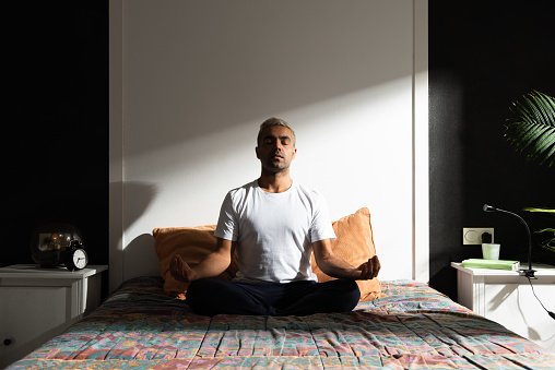 Hispanic man meditating in bedroom illuminated by sunlight. Healthy morning routine concept