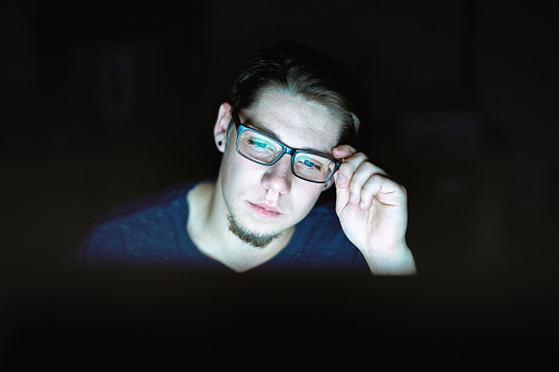Joven se sienta en la computadora, ajustándose las gafas, trabajando hasta tarde y cansado photo
