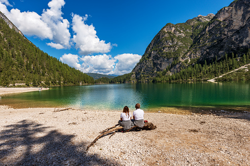 Lago di Braies, Italy - Sept 6th, 2021: Lago di Braies or Pragser Wildsee, small alpine beautiful lake and the Mountain peaks of the small and great Apostle (Piccolo Apostolo and Grande Apostolo), Dolomites, UNESCO world heritage site, Fanes-Senes-Braies nature park, South Tyrol, Trentino-Alto Adige, Bolzano province, Italy, Europe. Two adults, a man and a woman are relaxing, sitting on a tree trunk, on the shores of the beautiful lake on a sunny summer day.