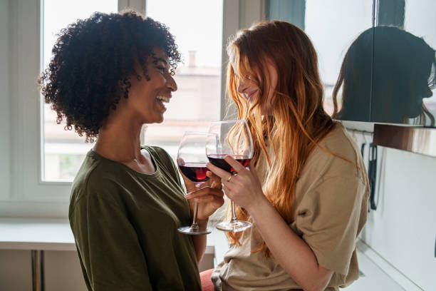 two happy homosexual girls, in intimacy with each other, enjoy a glass of wine in the kitchen, looking at each other and smiling together. stock photo