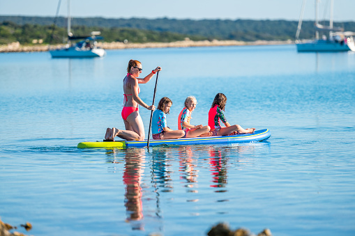 Happy Family on SUP stand up paddle on vacation. Active family riding SUP boards and paddling in the ocean on a beautiful morning. Mother and three girls share one SUP board with yachts in background