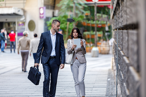 Attractive Business Couple is Walking in the City Streets and Talking about the Business Plans. Beautiful Businesswoman is Having a Conversation with a Handsome Business Partner While Walking in the City Center and Using a Tablet.