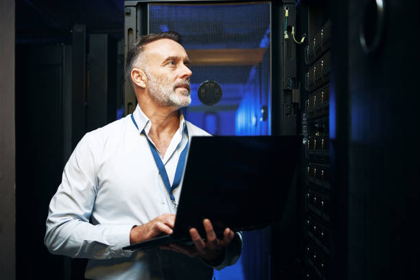 foto de un hombre maduro usando una computadora portátil mientras trabaja en una sala de servidores - computer programmer network server data center fotografías e imágenes de stock