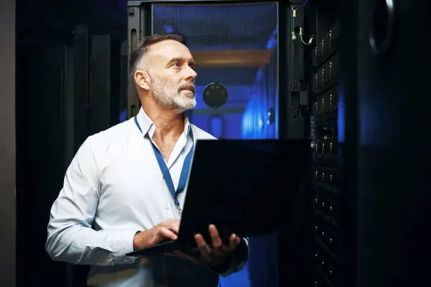 Photo of Shot of a mature man using a laptop while working in a server room