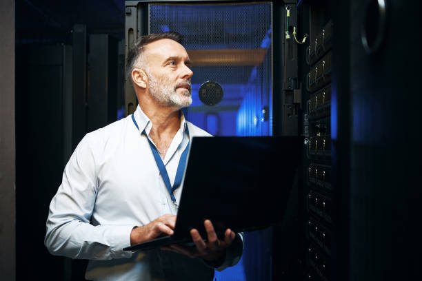 shot of a mature man using a laptop while working in a server room - computer network server repairing technology imagens e fotografias de stock