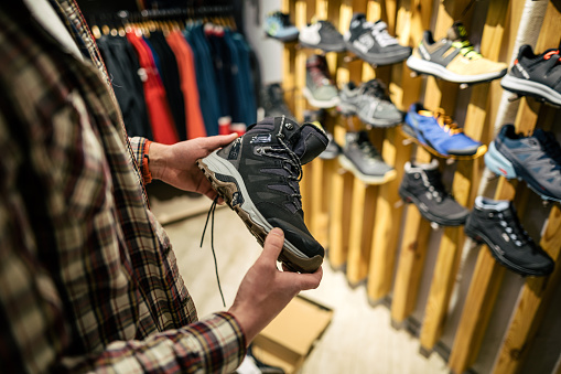 Caucasian man holds and examines hiking boots before buying in a shop for tourism and sports. Customer evaluates the quality of hiking shoes in a store. Waterproof hiking winter autumn boots.