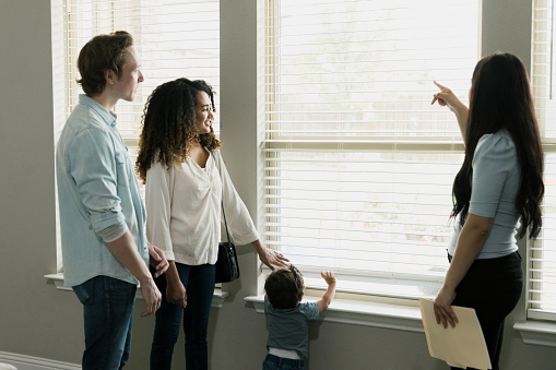 As their little boy plays with the blinds, the mid adult couple listens to the unrecognizable mid adult female real estate agent as she talks about the new home.