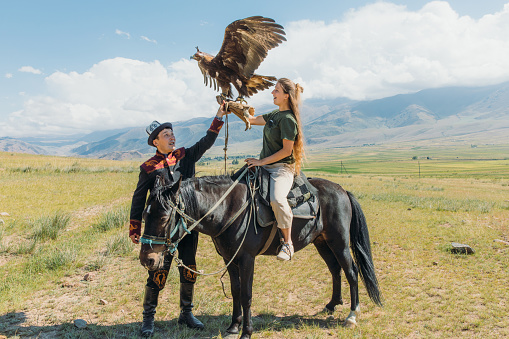 Young female tourist with long hair sitting on the horse holding Eagle and the hunter in traditional clothes near it, Tian Shan mountains, Kyrgyzstan, sunny summer morning at the plateau