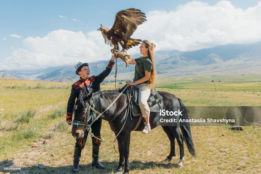 Woman traveler exploring the old traditions with Eagle Hunter in the mountains of Central Asia Young female tourist with long hair sitting on the horse holding Eagle and the hunter in traditional clothes near it, Tian Shan mountains, Kyrgyzstan, sunny summer morning at the plateau Hunter Stock Photo