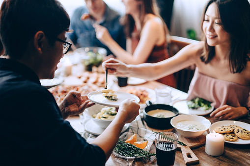 Joyful young Asian woman serving pasta to man. They are enjoying together, having fun, chatting and feasting on food and drinks at party