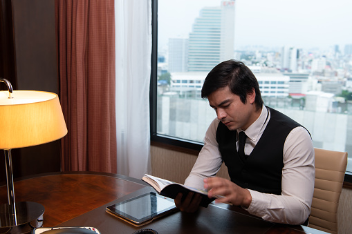 A young businessman monitors and keeps track of tasks in his office with a cityscape in the background of his desk.