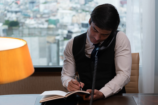 A young businessman monitors and keeps track of tasks in his office with a cityscape in the background of his desk.