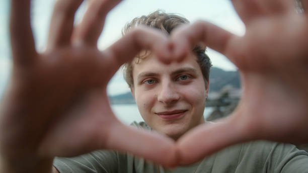 istock Young blond boy towards camera making heart symbol with hand and smiling looking at camera 1343562692