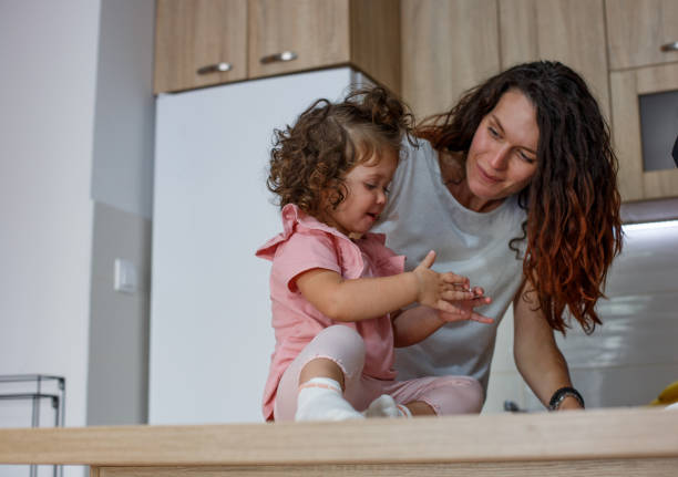 Little girl is playing with her mom in kitchen. Cute little girl is sitting in kitchen, on kitchen counter and playing with her mom. She is going to "help" her granny and make some holiday cookies. Her mom and her are smiling and enjoying in New Year day. baby new years eve new years day new year stock pictures, royalty-free photos & images