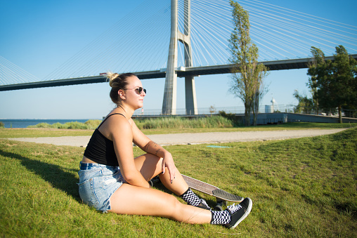 Pretty young woman with skateboard. Beautiful blonde girl in sunglasses with longboard sitting on grass near bridge, looking forward, relaxing. Sport, hobby, active lifestyle concept