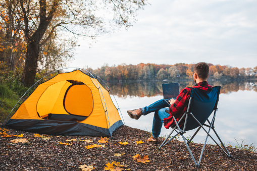 man sitting in chair near autumn lake working on laptop freelance camping concept