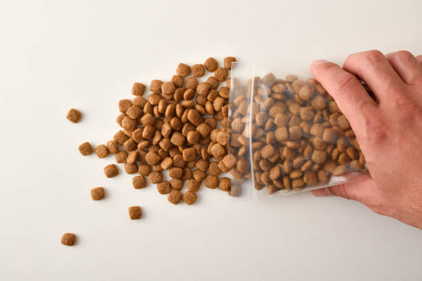 Man's hand dropping dog kibble from a clear plastic bag onto a white table. Top view. Horizontal composition.