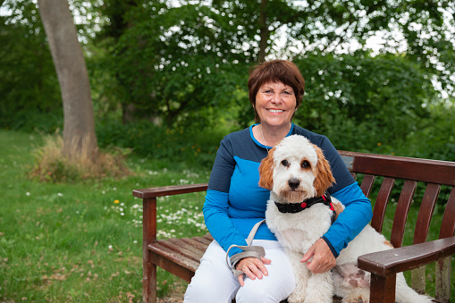 A caucasian senior woman wearing sports clothing on a summers day. She is looking and smiling at the camera while she sits on a bench on a dog walk with her Cavapoo dog.