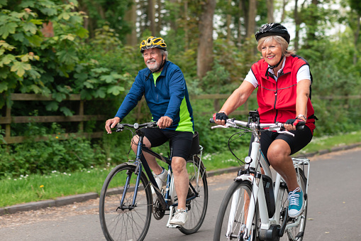 A caucasian senior couple wearing sports clothing and cycling helmets on a summers day. They are cycling down a road in a rural village.