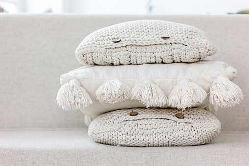 A pile of white and beige knitted sofa pillows with buttons and cotton tassels. Cozy textile textures, interior hygge and home handmade design concept