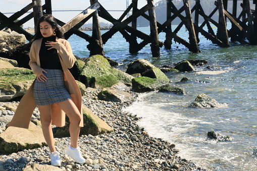 A model on a beach by the Pacific Ocean by a pier. She is wearing a black sweater, plaid mini skirt, white shoes and an open brown coat.