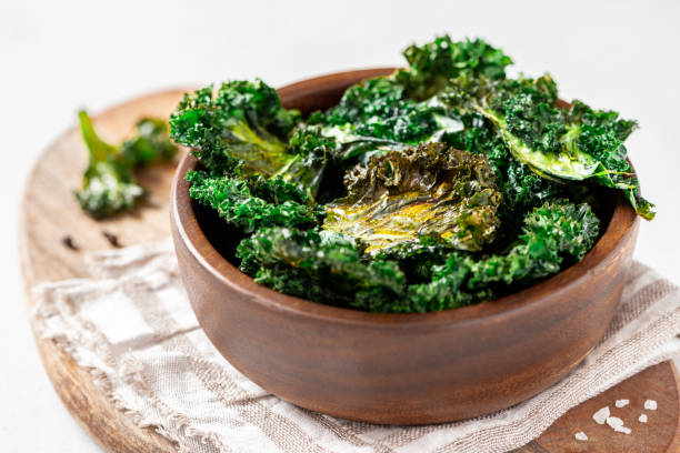 Baked kale chips in a wooden bowl Baked kale chips in a wooden bowl on a light background. Chips from the leaves of kale cabbage close-up. Kale chips with olive oil and salt, a tasty vegan snack. kale stock pictures, royalty-free photos & images