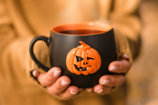 Black mug with a hot tea in female hands close-up.The mug is decorated with a pumpkin jack o'lantern.Halloween concept. stock photo
